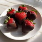Six perfect Chocolate Dipped Strawberry Hearts arranged on a white plate, showing the bright red fruit above the dark chocolate coating.