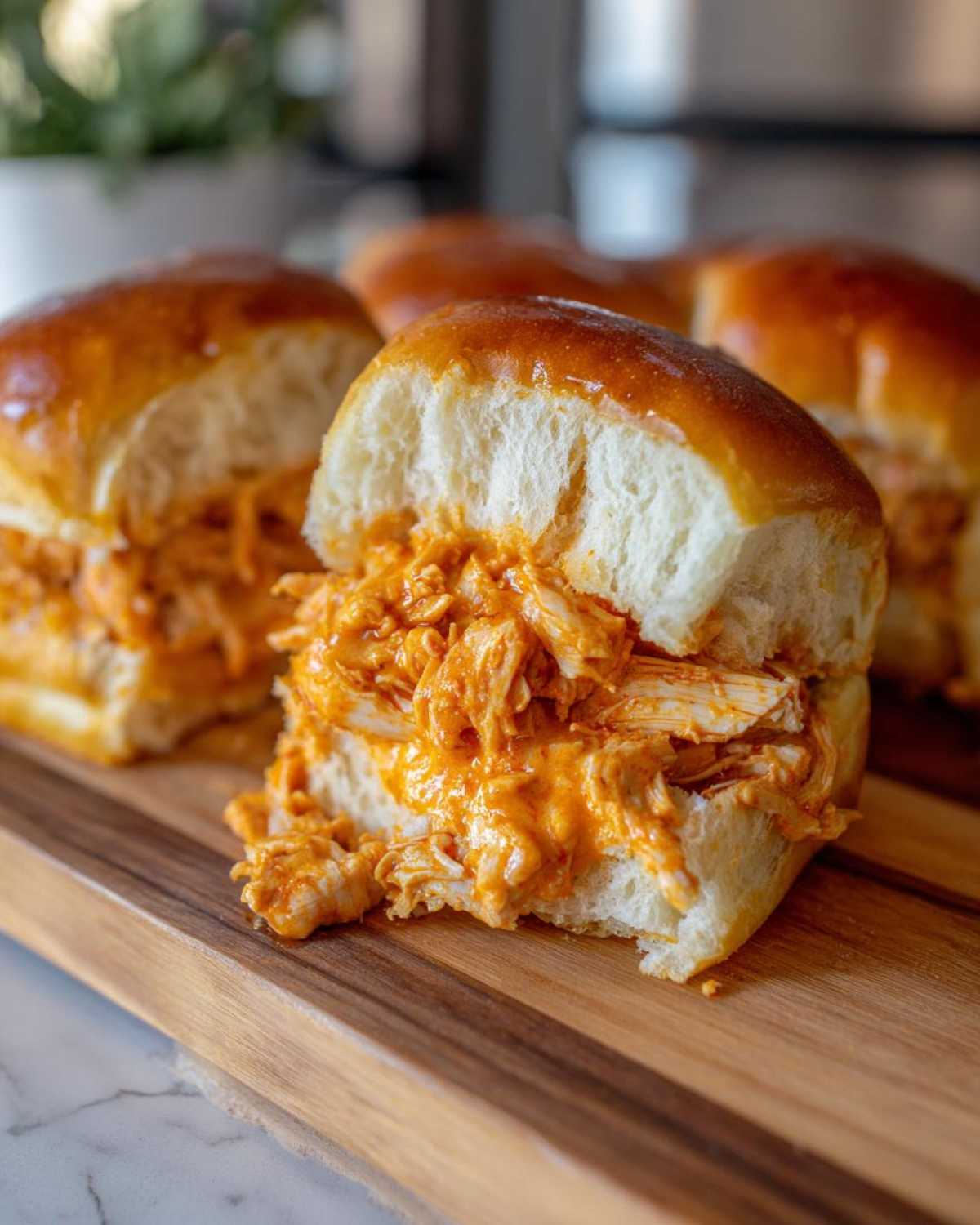 A close-up of a delicious Buffalo Chicken Sliders sandwich cut in half, showing shredded chicken mixed with orange sauce on a soft, glossy bun.