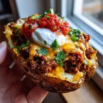 A hand holding one perfectly baked Taco Stuffed Potato Skins loaded with seasoned meat, melted cheese, sour cream, and salsa.