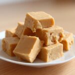 A stack of creamy, light brown peanut butter fudge cubes piled on a small white plate.
