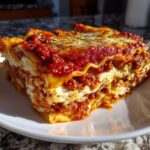 A close-up, appetizing slice of 9 layer lasagna showing layers of pasta, ricotta, and rich meat sauce.