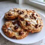 Close-up of three freshly baked almond joy cookies loaded with chocolate chips, coconut flakes, and nuts.