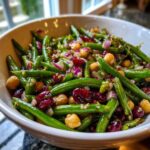 A close-up view of a vibrant bean salad featuring bright green beans, chickpeas, dried cranberries, and red onion.