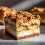 A close-up, cross-section view of one apple cheesecake bars showing layers of crust, creamy filling, spiced apples, and crumble topping.