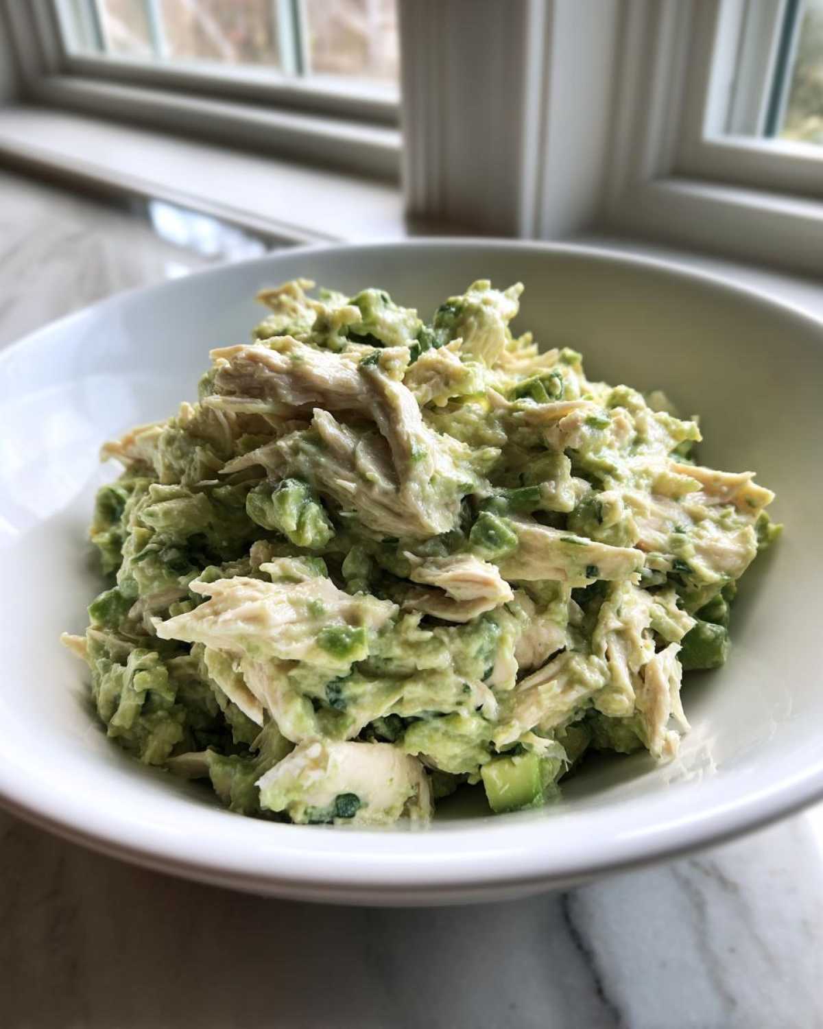 A close-up view of creamy avocado chicken salad, featuring shredded chicken mixed with mashed avocado and green onions, served in a white bowl.