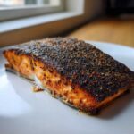 Close-up of a thick fillet of blackened salmon with a dark, spicy crust resting on a white plate.