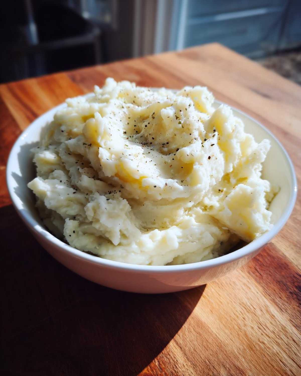 A close-up of creamy Boursin mashed potatoes seasoned with pepper, served in a white bowl on a wooden table.