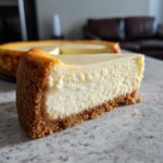 A thick slice of creamy baked cheesecake with a graham cracker crust, part of the whole cheesecake visible behind it.
