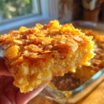 A hand holding a square serving of cheesy potato casserole with a golden, crunchy corn flake topping.