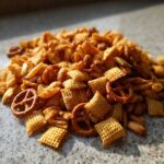 A close-up pile of freshly seasoned chex party mix featuring square cereal pieces and small pretzels.