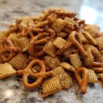 A mound of seasoned chex party mix featuring square Chex cereal and small pretzels piled on a granite countertop.