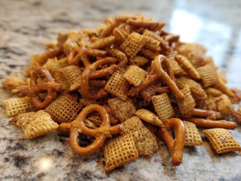 A mound of seasoned chex party mix featuring square Chex cereal and small pretzels piled on a granite countertop.