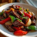 Close-up of glossy, dark brown slices of Chinese pepper steak mixed with vibrant red and green bell peppers and onions.