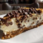 A close-up of a rich slice of chocolate chip cheesecake on a white plate, featuring a graham cracker crust and chocolate chips throughout.