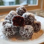A close-up of several dark chocolate bourbon balls stacked on a white plate, heavily coated in shredded coconut.