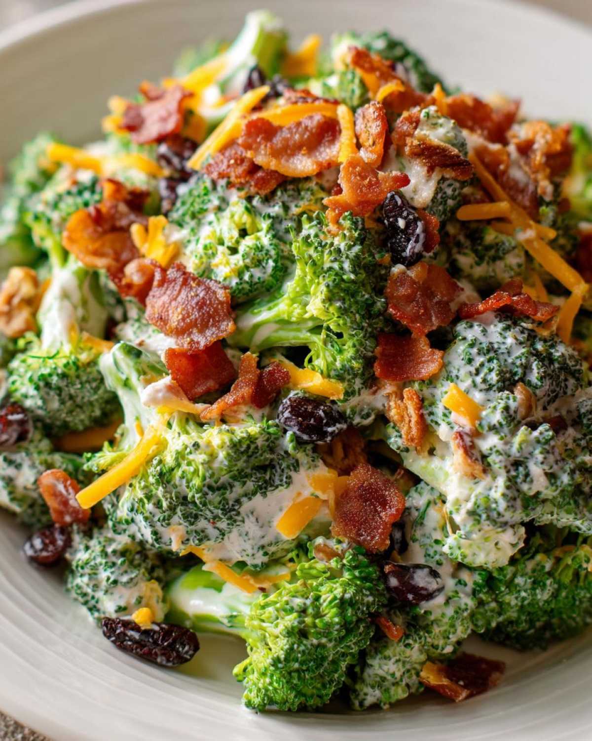 A close-up view of a creamy broccoli salad topped generously with crispy bacon bits, shredded cheddar cheese, and raisins.