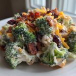A close-up serving of creamy broccoli salad featuring florets coated in dressing, topped with bacon bits, shredded cheddar, and dried cranberries.