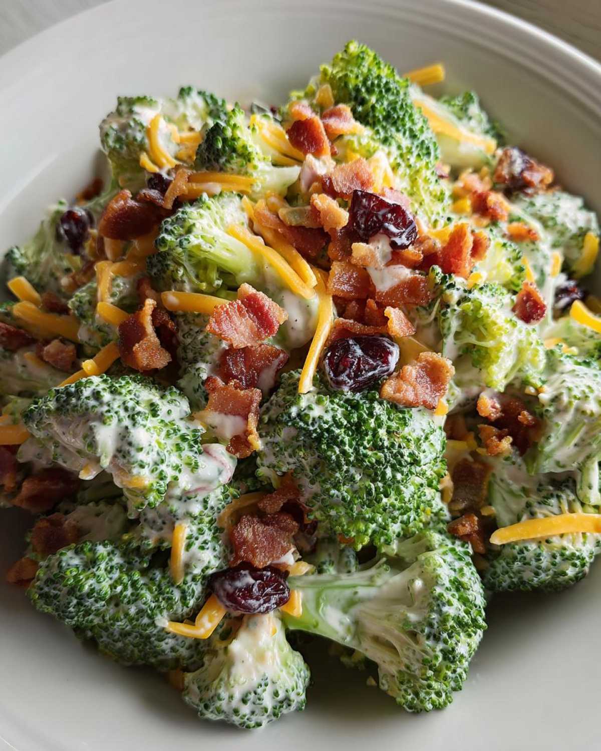 A close-up view of a creamy broccoli salad tossed with shredded cheddar cheese, bacon bits, and dried cranberries.