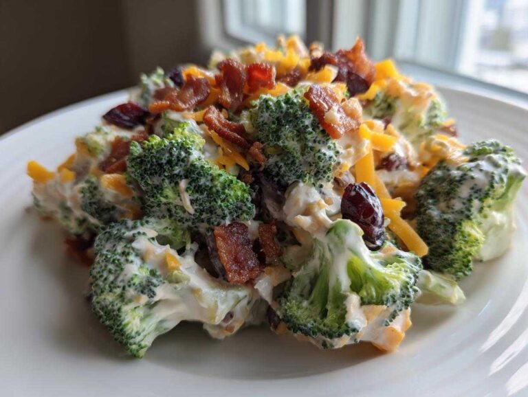 A close-up serving of creamy broccoli salad featuring florets coated in dressing, topped with bacon bits, shredded cheddar, and dried cranberries.