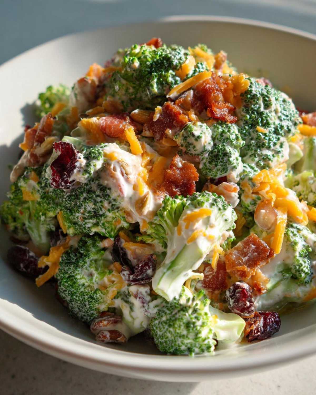 A close-up of a bowl filled with creamy broccoli salad, topped with cheddar cheese, bacon bits, and dried cranberries.