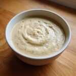 A close-up of a white bowl filled with thick, creamy cauliflower soup, topped with freshly ground black pepper.