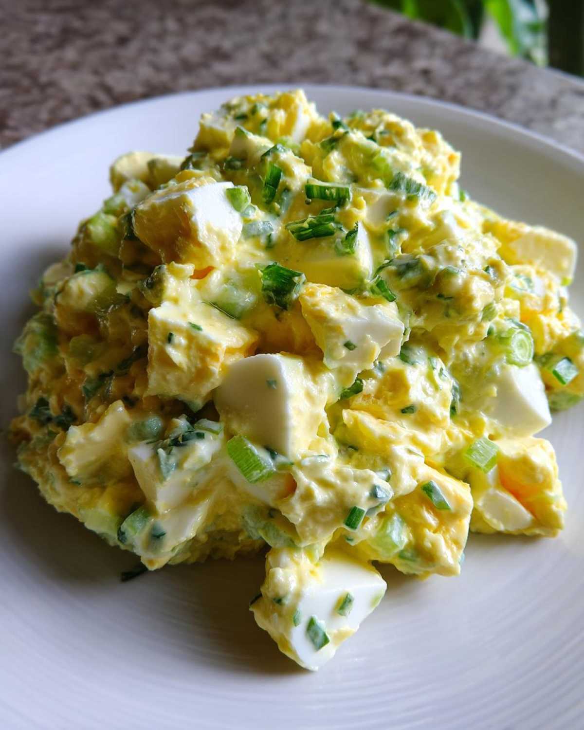 A mound of chunky, creamy egg salad recipe featuring visible chunks of white and yellow egg mixed with green chives on a white plate.