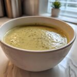 A close-up of a white bowl filled with creamy, pale green leek soup, seasoned with pepper.