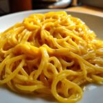 A close-up shot of bright yellow, glossy creamy lemon pasta tossed perfectly on a white plate, ready to eat.