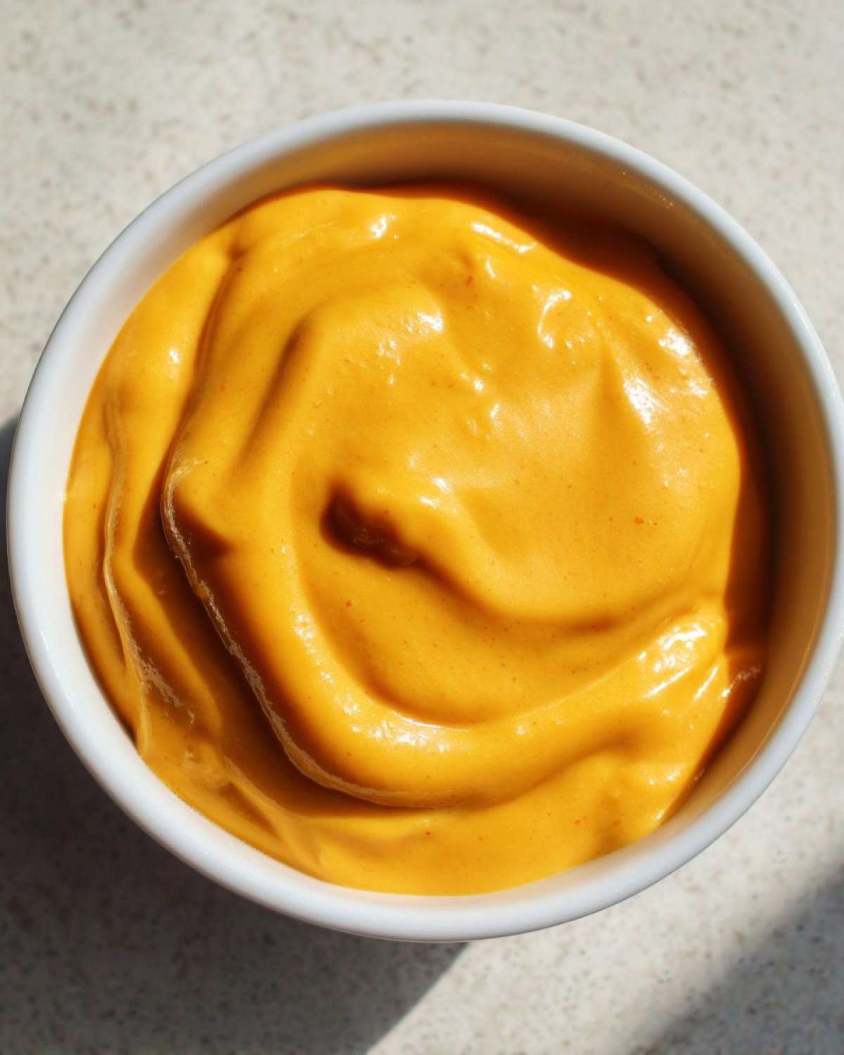 Close-up overhead shot of thick, vibrant orange nacho cheese sauce swirled inside a small white bowl.