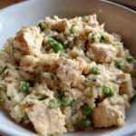 Close-up of creamy one pot chicken and rice mixed with bright green peas and herbs in a white bowl.