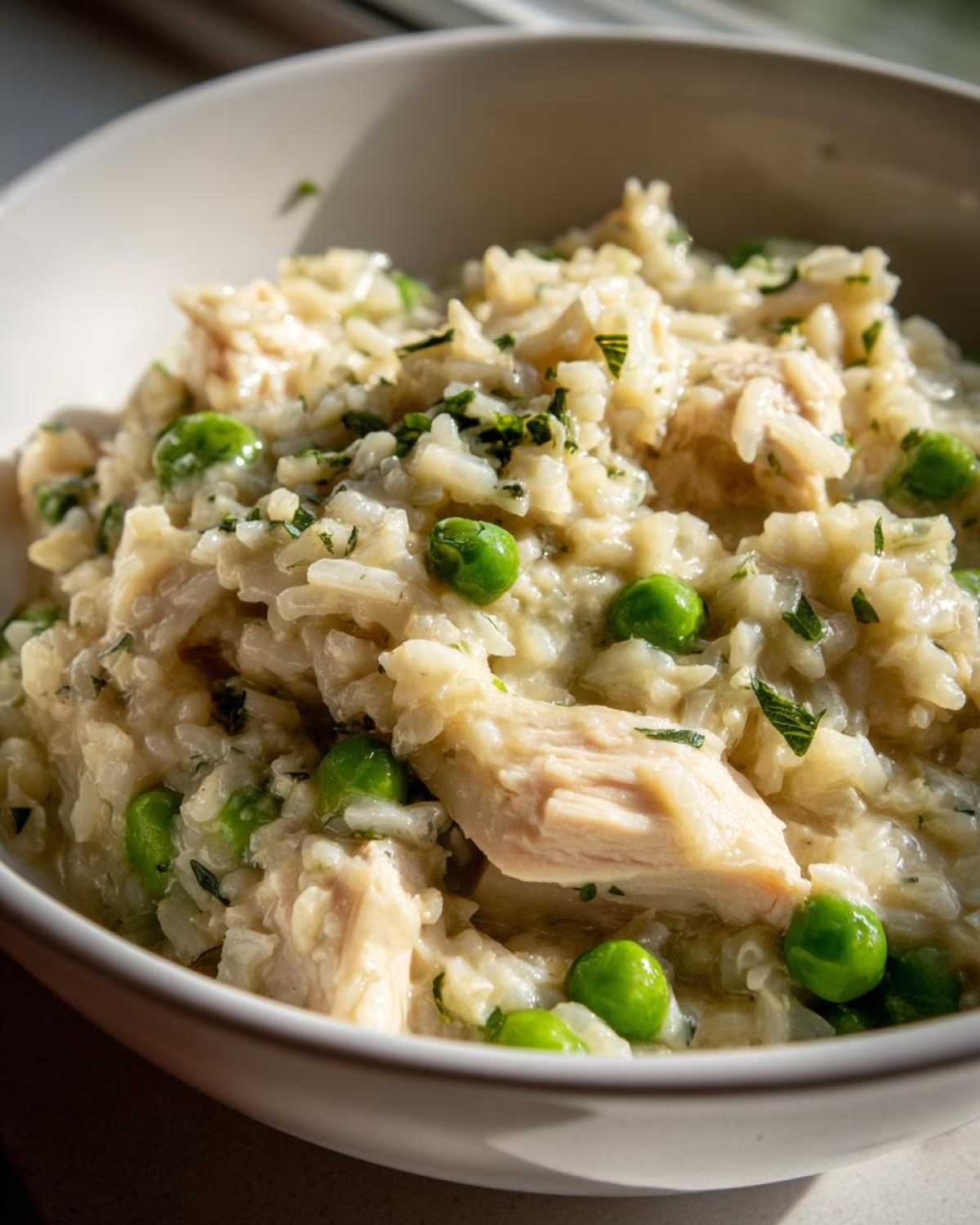 Close-up of creamy one pot chicken and rice mixed with bright green peas and herbs in a white bowl.