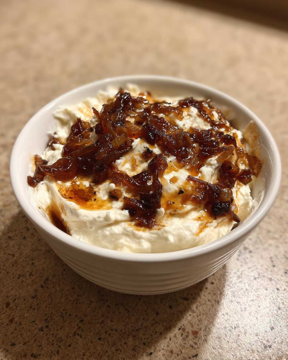 A white bowl filled with creamy onion dip, generously topped with rich, dark caramelized onions.