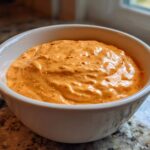 Close-up of a creamy, orange-hued rotel dip speckled with red chili flakes in a white serving bowl.