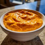 Close-up of creamy, bright orange rotel dip served in a white bowl, showing texture and flecks of red.