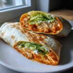 A crispy buffalo chicken wrap cut in half, showing shredded chicken coated in orange sauce, lettuce, and ranch dressing.