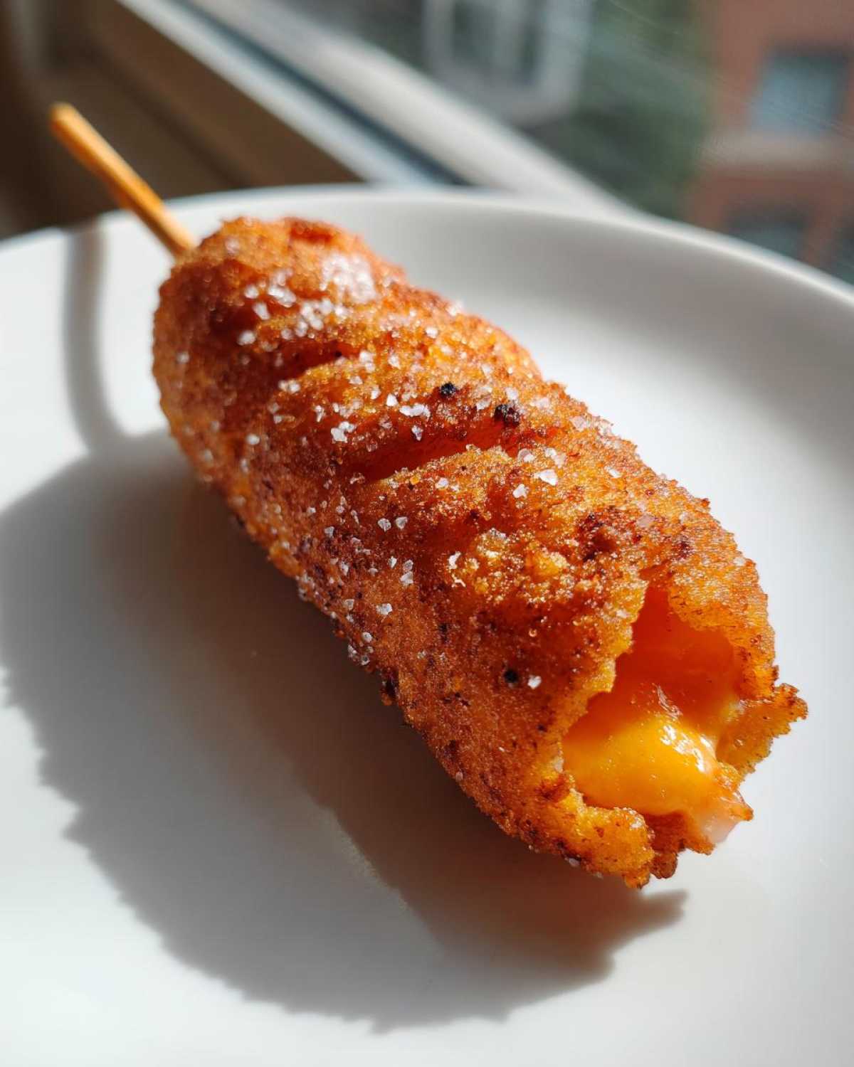 Close-up of a crispy, sugar-dusted korean corn dog on a stick, showing melted cheese oozing from the end.