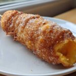 A close-up of a freshly fried korean corn dog on a stick, heavily coated in granulated sugar.