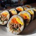 A row of freshly sliced tuna kimbap rolls showing seasoned rice, ground tuna filling, julienned carrots, and egg strips wrapped in seaweed.