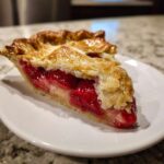 Close-up of a slice of strawberry rhubarb pie showing juicy red filling and a golden, flaky top crust.