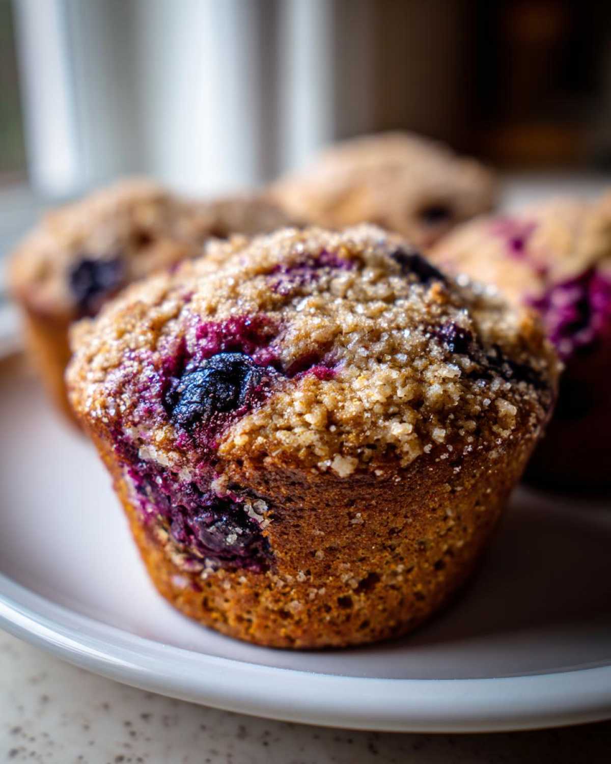 A close-up of a fluffy blueberry oatmeal muffin topped with brown sugar streusel, sitting on a white plate.
