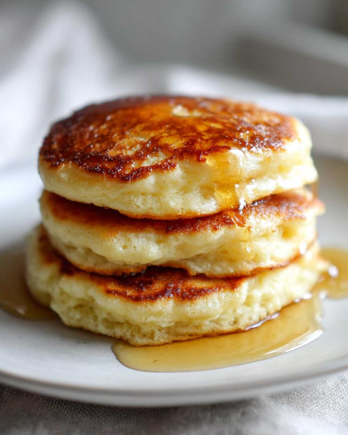 Close-up of a stack of three fluffy greek yogurt pancakes drizzled with golden syrup on a white plate.