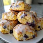 A stack of fluffy lemon blueberry scones generously topped with a white lemon glaze, served on a white plate.