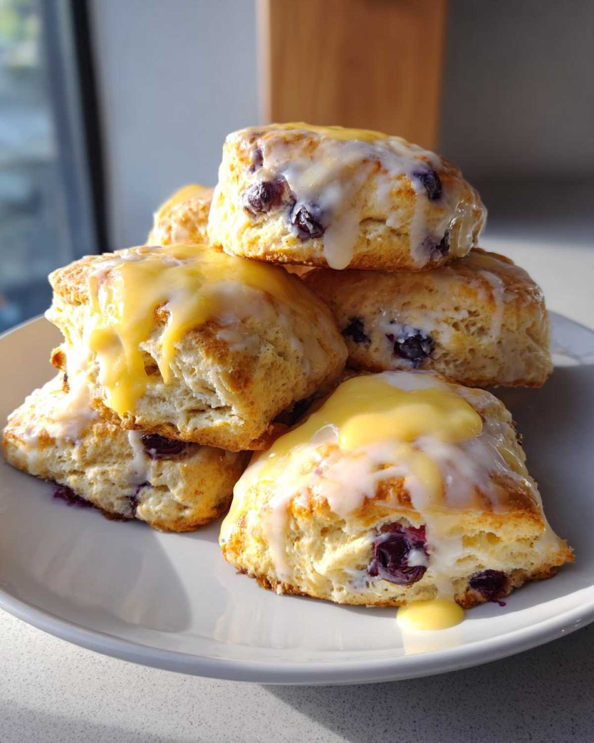 A stack of fluffy lemon blueberry scones topped with a thick lemon glaze, sitting on a white plate.