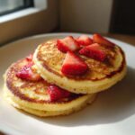 Two fluffy strawberry pancakes stacked on a white plate, topped with fresh sliced strawberries.