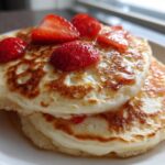 Close-up of two fluffy strawberry pancakes stacked, topped with fresh strawberries and drizzled with syrup.