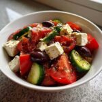Close-up of a vibrant greek salad featuring large chunks of tomato, cucumber, feta cheese, and Kalamata olives, dressed with herbs.