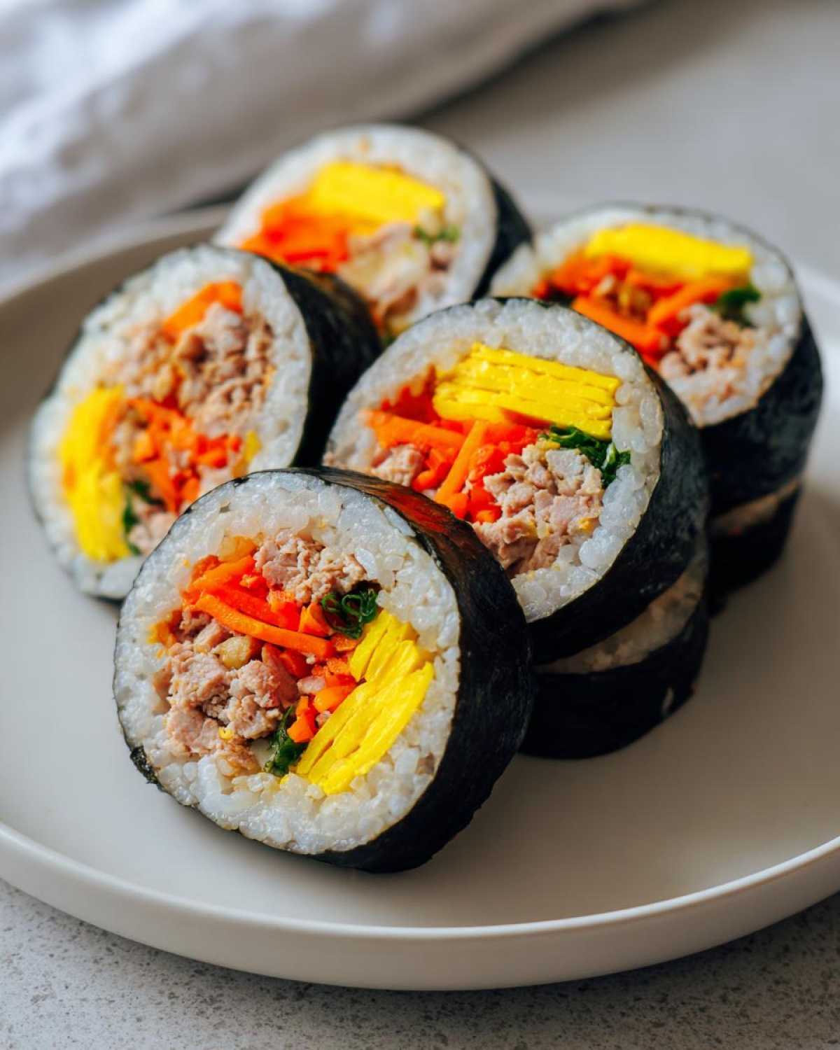 Five slices of vibrant tuna kimbap, showing fillings of tuna, julienned carrots, and yellow egg strips, arranged on a white plate.