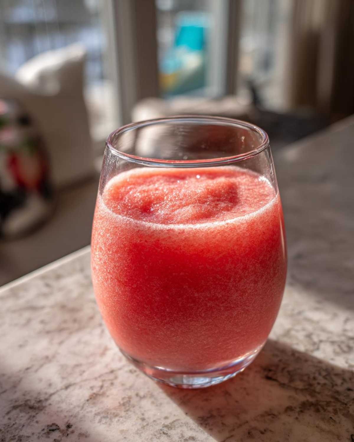 A close-up of a vibrant, frozen strawberry daiquiri served in a clear glass on a marble countertop.