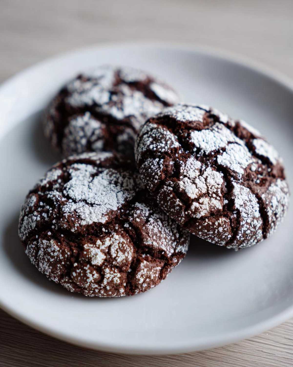 Close-up of three perfectly baked chocolate crinkle cookies dusted heavily with powdered sugar on a white plate.