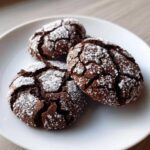 Three fudgy chocolate crinkle cookies heavily dusted with white powdered sugar on a white plate.